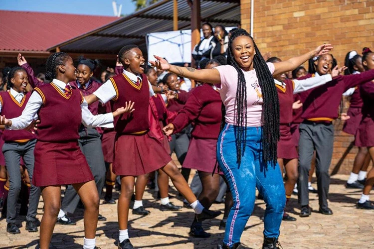 A black woman, with long dark braids, dances with a happy expression on her face. She guides a large group of black adolescent school girls in dark red school uniforms.