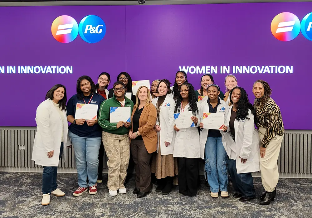 A group of women smile and pose, with some holding white certificates in their hands. Some of them wear white lab coats. Behind them, a purple screen says, “Women In Innovation” and features a round, blue P&G logo in the upper right corner.