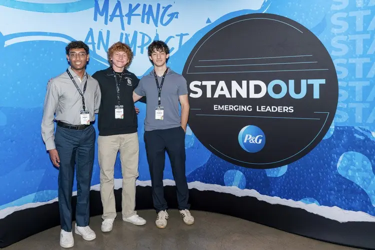 Three male students wearing event badges stand together beside a large black and blue sign that says “Standout Emerging Leaders.”