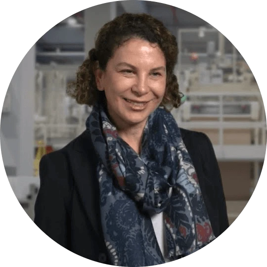 A woman with short curly haired. She wears a dark jacket and scarf and stands inside a manufacturing plant.