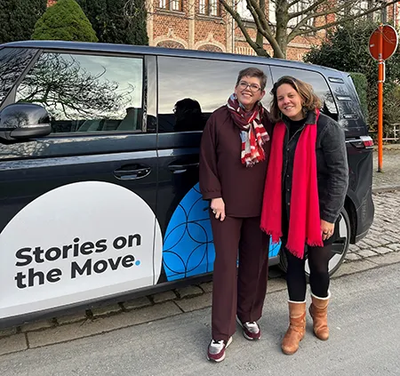 Two women in dark clothes and warmly colored scarves stand together in front of a dark van with a white and blue circular logo on the door that says, “Stories on the Move.”