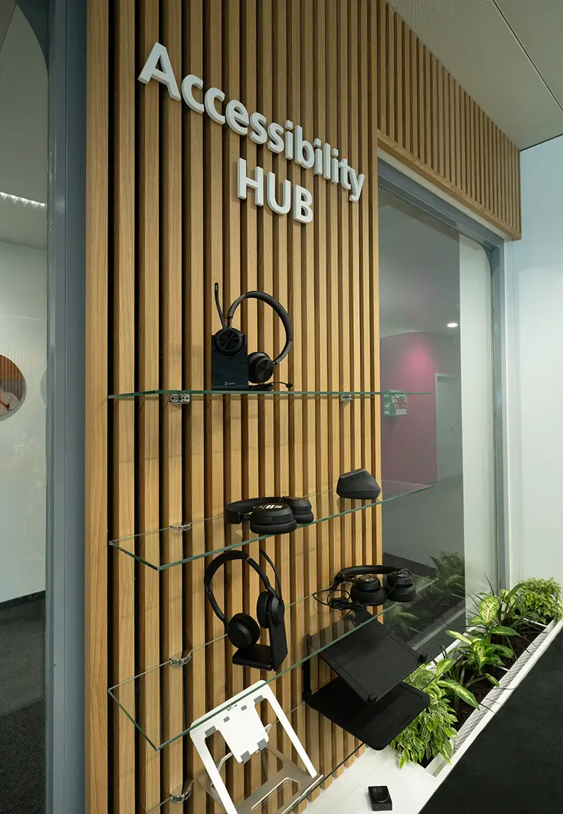 Wooden paneled wall with glass shelves displaying headsets, headphones, a tablet stand, and other assistive tech devices.