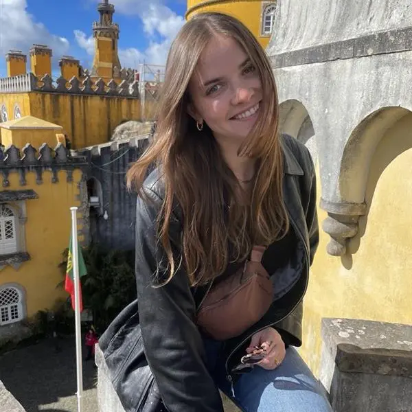 A young woman with long blonde hair, jeans and a black leather jacket. She stands in front of a large yellow castle.