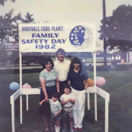 A mother and father pose with their teen daughter and two small children. They stand next to a white blue sign that says “Ivorydale food plant. Family safety day 1982.”
