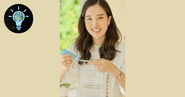 An Asian woman with long dark hair wears a white embroidered blouse. In her hands, she holds up a blue tube of oral care product and a white applicator, while she smiles directly into the camera.