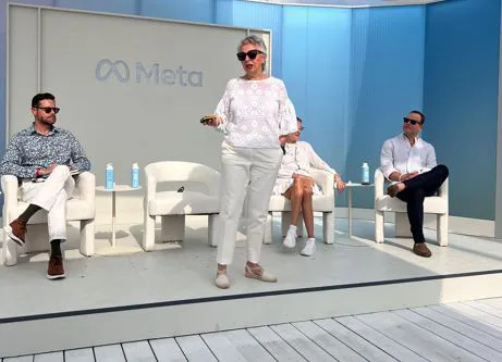 Two men and one woman sit on a stage with a backdrop panel that reads ‘Meta’ as a woman with sunglasses on stands and speaks.