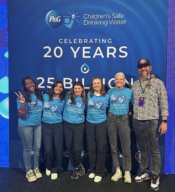 Five women and one man smile and stand together in front of a large blue post. Most of the group is wearing matching light blue t-shirts.