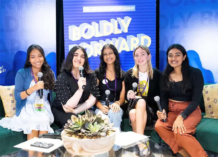 Five girls of different ethnicities smile while seated on a green chair, surrounded by yellow pillows as, they hold microphones at the Girl Up Leadership Summit. A blue background displayed a white tagline that reads, “Boldly Forward.”