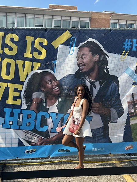 A young woman in a white dress and red purse poses in front of a large banner featuring two smiling individuals and the text "THIS IS HBCU."