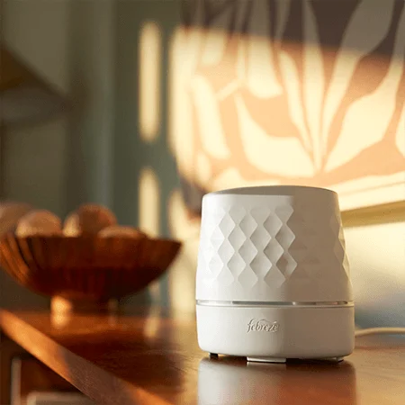 A white oval shaped device sits on a brown table. A decorative basket with balls sits at the far end. Sunlight hits the framed art in the background.