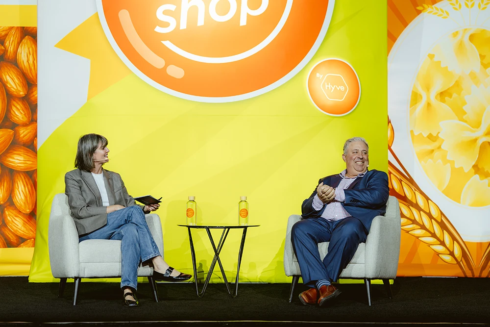 A woman and a man sit in armchairs on a stage, engaging in a discussion. The backdrop features a "shop by Hyve" logo and food illustrations.