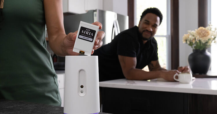 Two people in a kitchen. A person in the foreground places a small white airia scent cartridge into a white airiaI scent diffuser. Person in the background looks on.