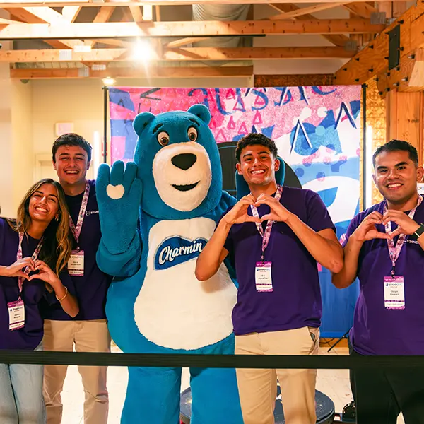 Three young men and a young woman in purple shirts smile. They are standing next to someone in a blue bear costume.
