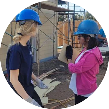 Two women wearing construction hats and gloves are on a construction site.