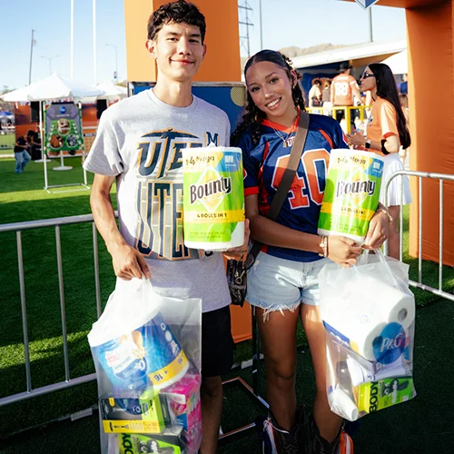 A young man and woman hold bags of everyday household products, including rolls of paper towels, in front of a booth at an outdoor event.