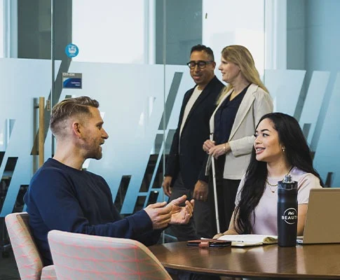 A man and woman are working together at a conference table, talking together. Another man and woman are walking outside the glass-walled room, and another man works at a separate table, his back to the others.