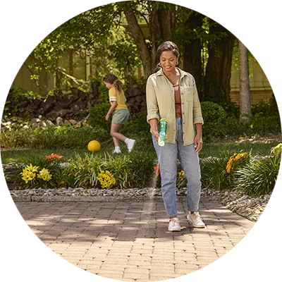 A black woman in jeans and a light brown shirt stands in her yard. She sprays a liquid on a weed.