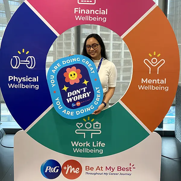 A young Asian woman in a white shirt and glasses. She stands behind a large, colorful round display and smiles.