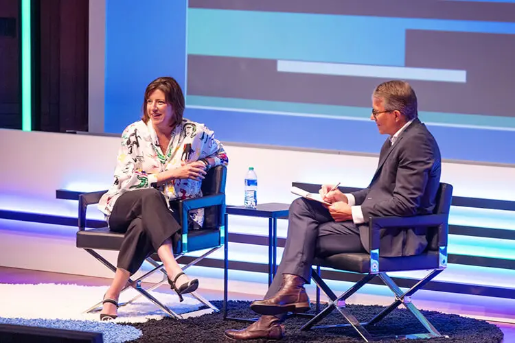 A woman with short brown hair wears a white patterned blouse, black pants and black heels. A man sits next to her, wearing a business suit. They are having a conversation on a stage.