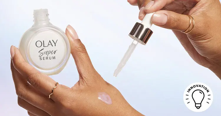 A woman's left hand holds a round olay super serum bottle between her thumb and index finger, as a small amount of iridescent serum is displayed on the side of her hand. Her right hand hovers above her left hand as it holds the bottle's dropper.
