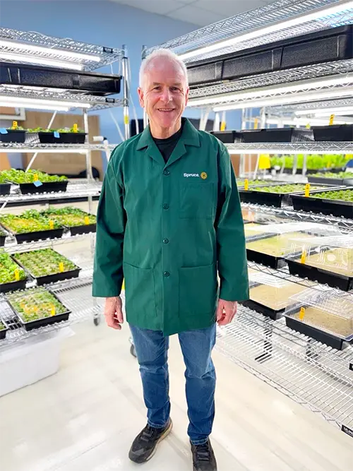 A smiling man in a green lab coat with a "Spruce" logo on the pocket, stands in an indoor plant cultivation facility. He is surrounded by multi-tiered shelving units holding trays of young plants under bright grow lights.