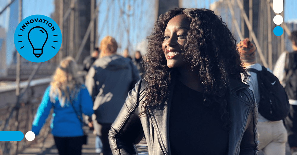 A black woman with long dark curly hair wears a black shirt and leather jacket. She smiles as she stands on the Brooklyn Bridge, looking off into the distance.