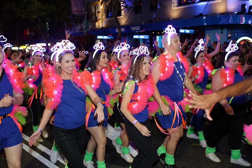 Over a dozen men and women smile as they march together in an evening parade. They are wearing matching outfits of blue shorts and shirts, pink feathered scarves and battery lit necklaces and crowns.