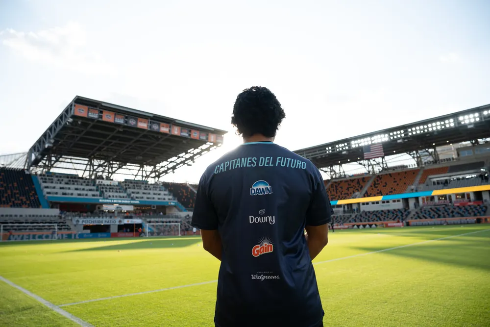 A young man in a uniform with logos of P&G brands on the back stands facing a soccer field.