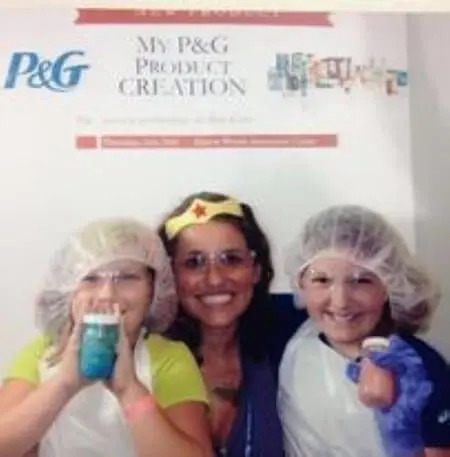 A woman hugs her two daughters. They are wearing aprons and hair nets and are standing in front of a Procter and Gamble product creation sign.