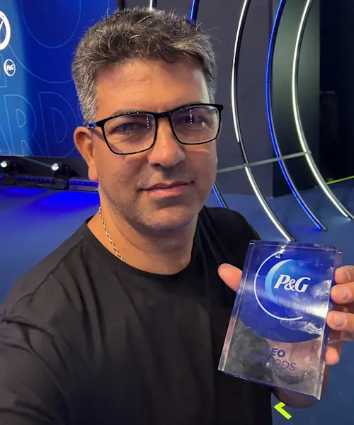 A man with grey hair and black glasses holds up a P&G award, with blue and white lights in the background.