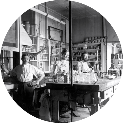 Two white men in white shirts and dark pants and one white woman in a white shirt and a dark skirt stand inside an industrial laboratory. In the black and white photo, they are surrounded by lab flasks and other materials.