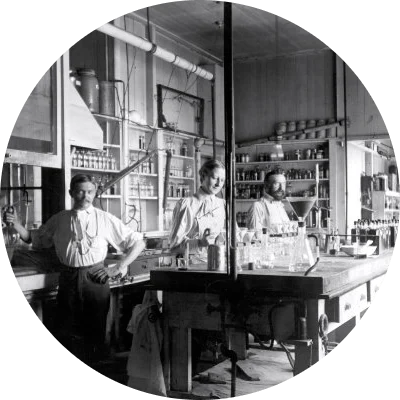 Two white men in white shirts and dark pants and one white woman in a white shirt and a dark skirt stand inside an industrial laboratory. In the black and white photo, they are surrounded by lab flasks and other materials.