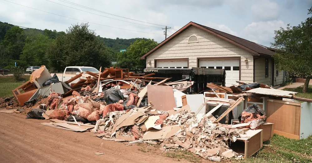 A large pile of flood-damaged debris, including drywall, insulation, cabinets, and household materials, sits outside a home along a muddy roadside.