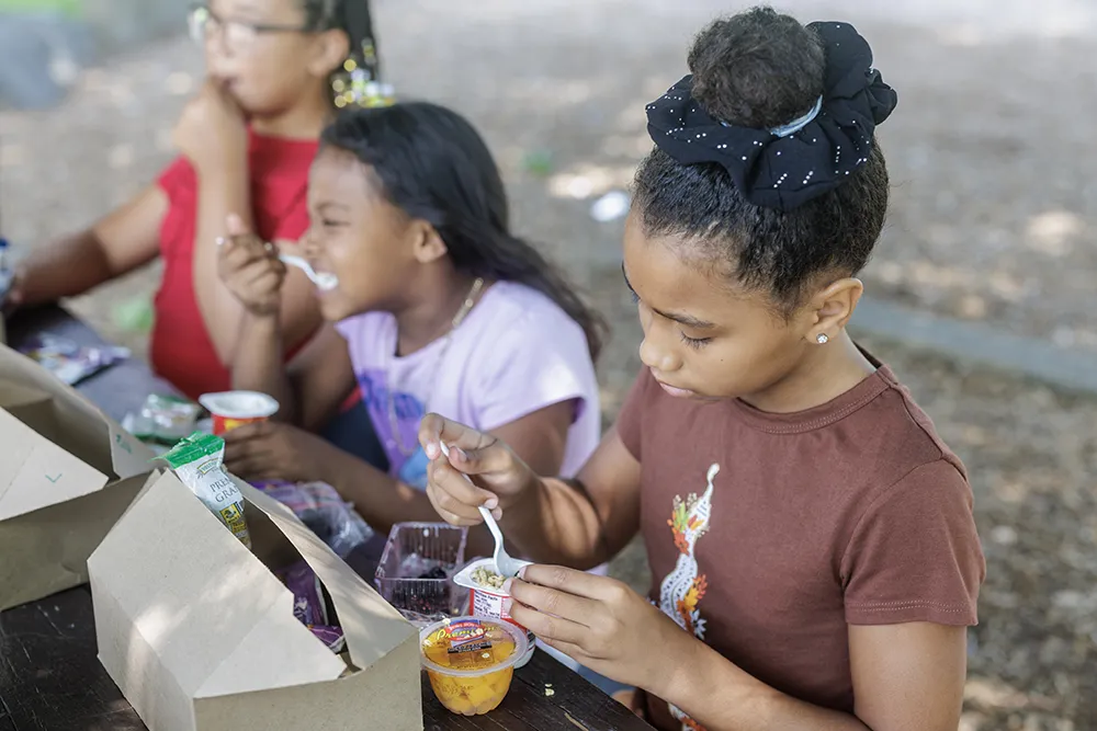 Three young girls sit at an outdoor picnic table enjoying a packed meal, with one girl in a brown shirt opening a small container of food.