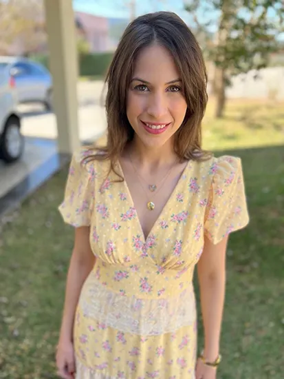 A woman with brown shoulder-length hair. She wears a yellow dress and stands in her yard with a tree and vehicles in the background.