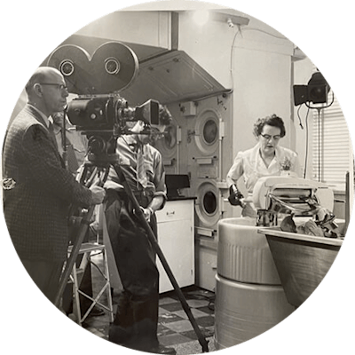 A woman in a white uniform operates a mid-century mechanical laundry machine. Two men stand behind a camera tripod as they film her doing laundry. The image is in black and white.