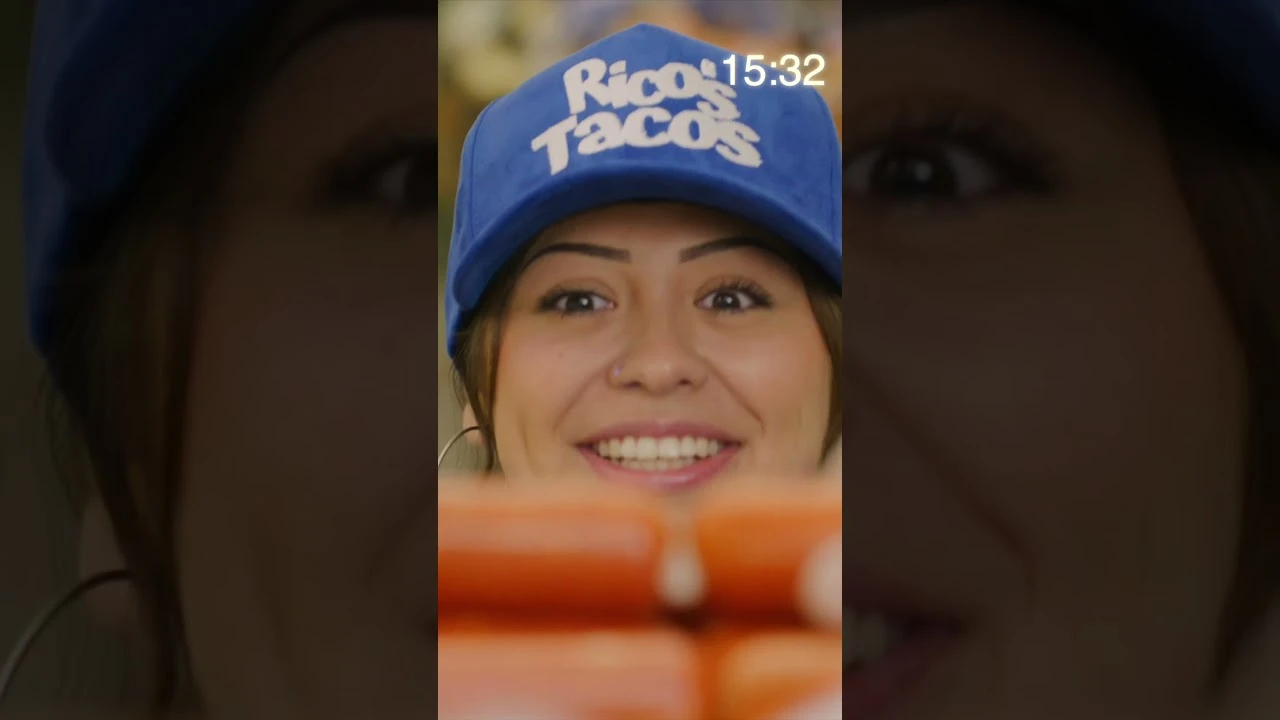 Close-up of a restaurant worker wearing a Rico’s Tacos cap, looking toward the camera behind a food counter.