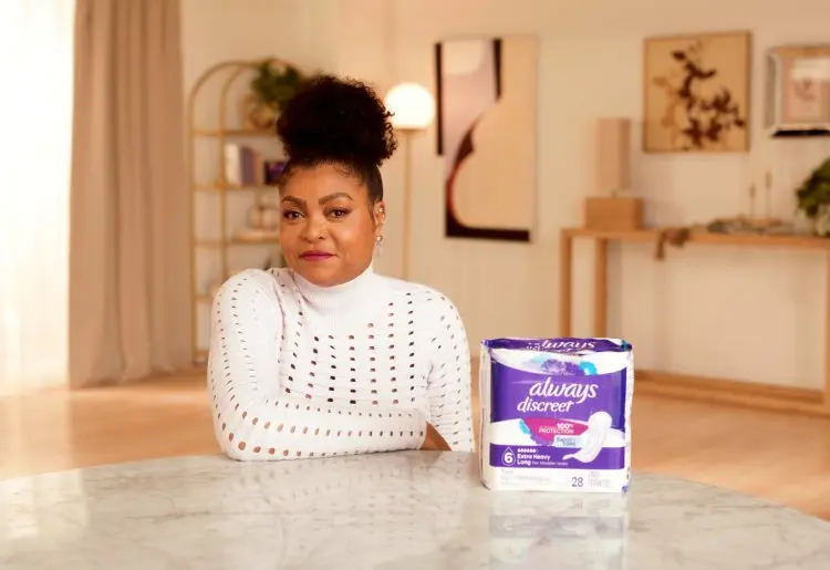 A black woman wears a white shirt and smiles as she sits at a dining table. A white and purple plastic package of Always Discreet feminine pads sits on the table next to her.
