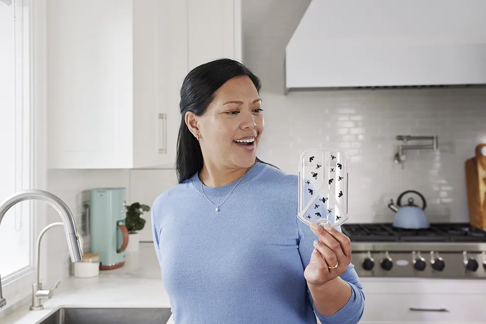 An Asian woman stands in a kitchen. She holds up a clear cartridge for an insect and bug device.