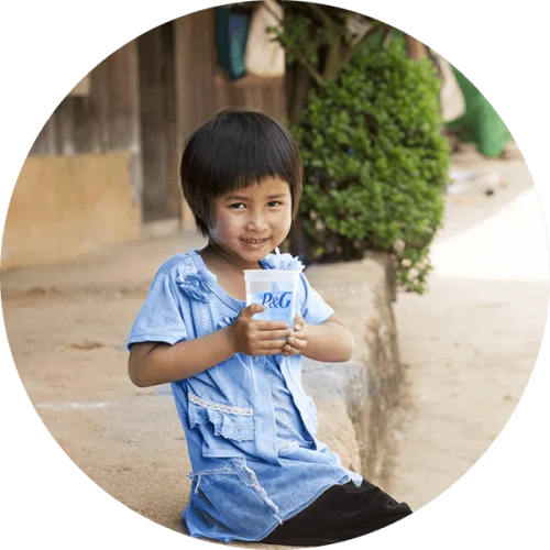 A young girl with black hair in a blue shirt sits outside smiling, holding a P&G cup of clean water.