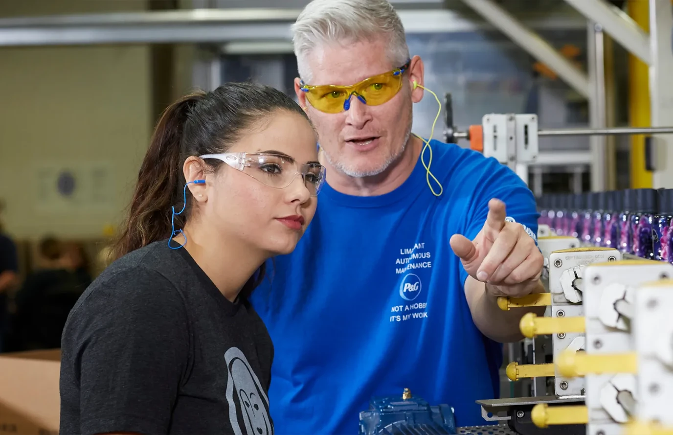 Two colleagues in a lab—man in blue shirt explaining to woman in dark blue; both wear safety glasses.