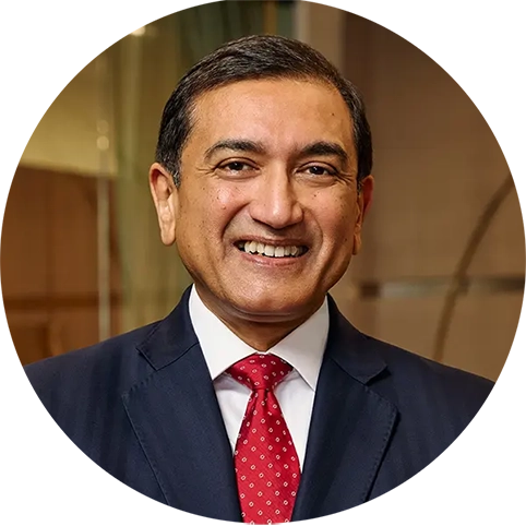 Professional headshot of Shailesh Jejurikar wearing a dark suit, white shirt, and red tie, facing the camera in an office setting.