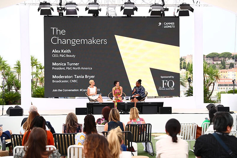 One black woman and two white women dressed in business dresses sit together on an outdoor stage, while rows of people in the audience watch.