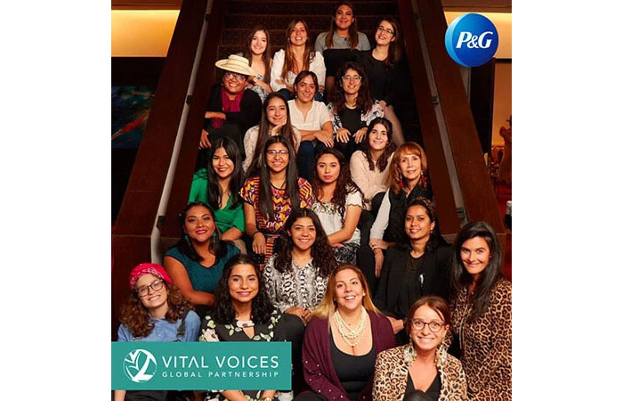 A large group of women and girls pose together on a staircase and smile. A white and teal logo in the bottom left corner says, "Vital Voices." In the top right corner, there is a round, blue logo with the word "P&G" on it.