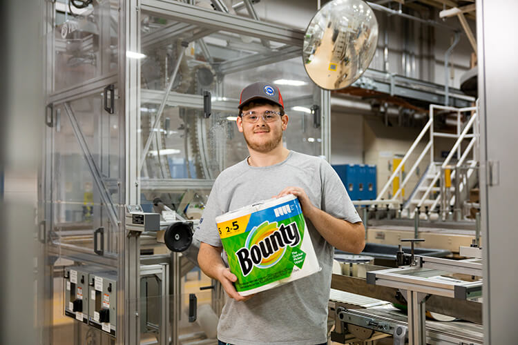 A man in a grey cap and grey t-shirt stands in a manufacturing plant as he holds a green plastic pack of Bounty paper towels.