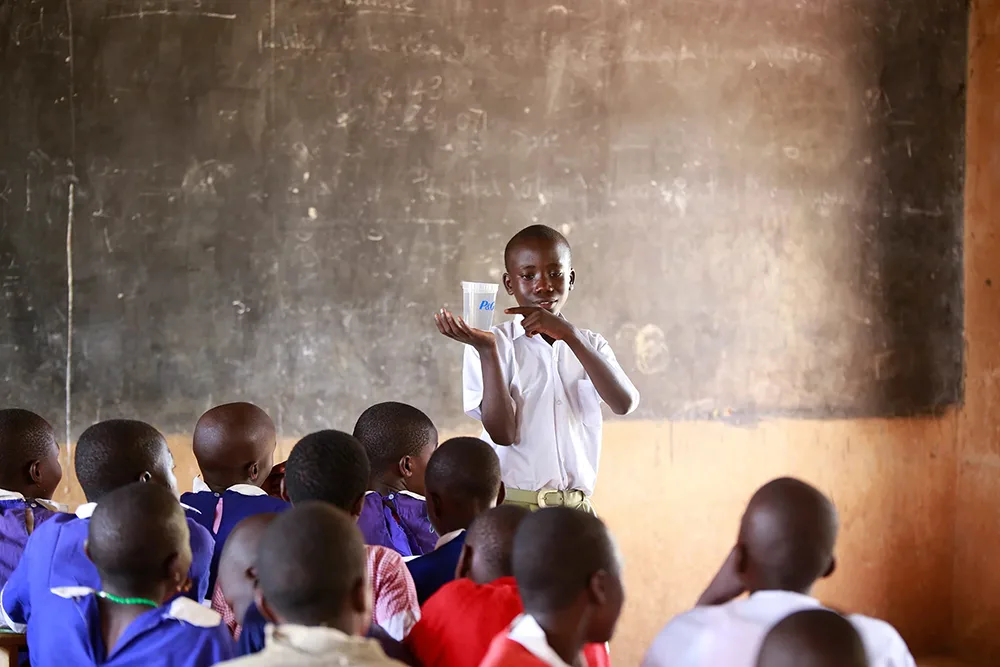 A young African boy in a white shirt stands at the front of a classroom holding up a clear cup, pointing to it as other children sit facing him against a worn chalkboard backdrop.