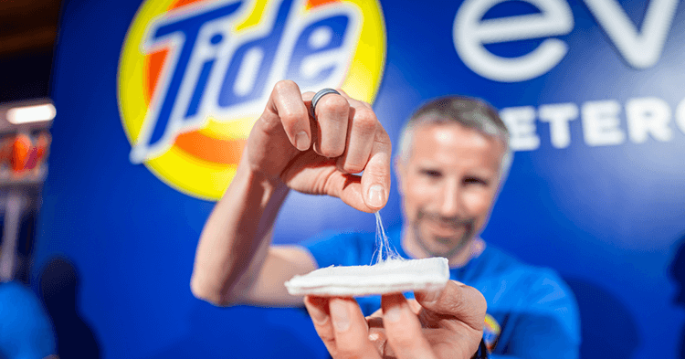 A man holds up a white square shaped swatch in one hand. With the other he is pinching and lifting white fibers from the swatch. The round yellow, orange and blue Tide logo is displayed on a bright blue backdrop.