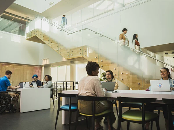 Several people sit together at various office work stations. Two women walk down a staircase in the background.
