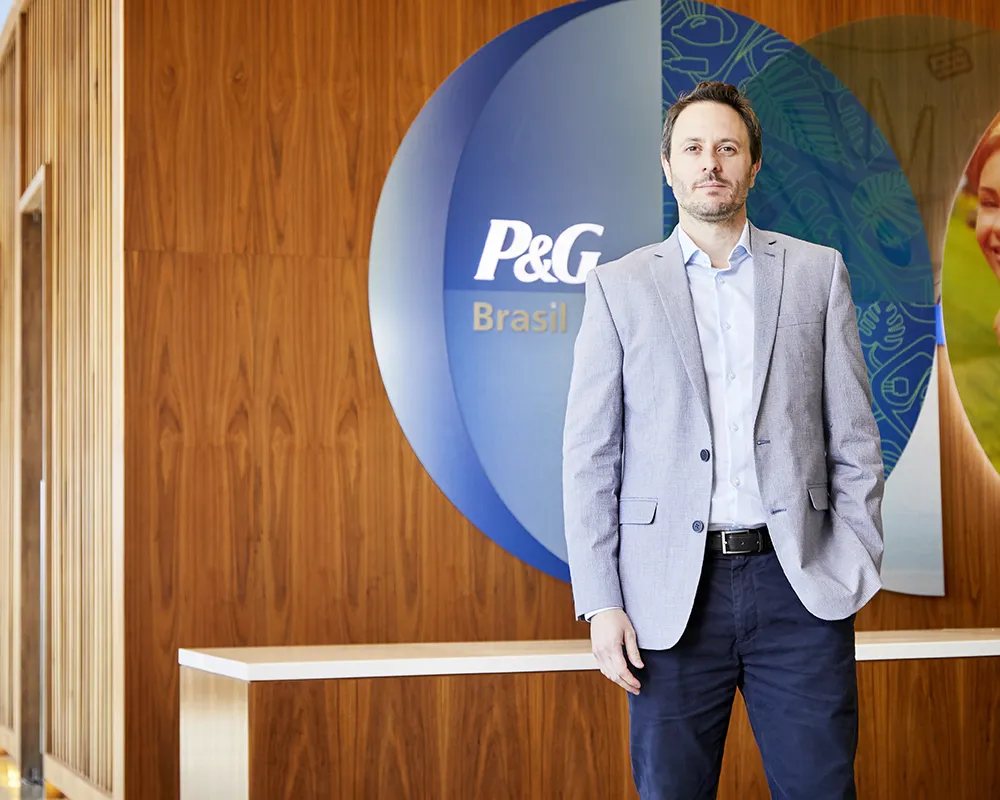 A man in jeans, grey jacket and white collared shirt. A P&G Brazil logo hangs on a wall behind him.