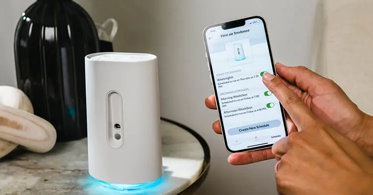 A close up of a person’s iPhone displays the airia app. A white airia scent diffuser is placed next to the phone on a night stand.